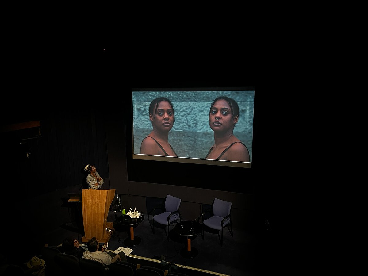 Jasmine standing at a lectern in front of a screen showing a still from the film Sugar Coated. On the screen there are two Jasmines