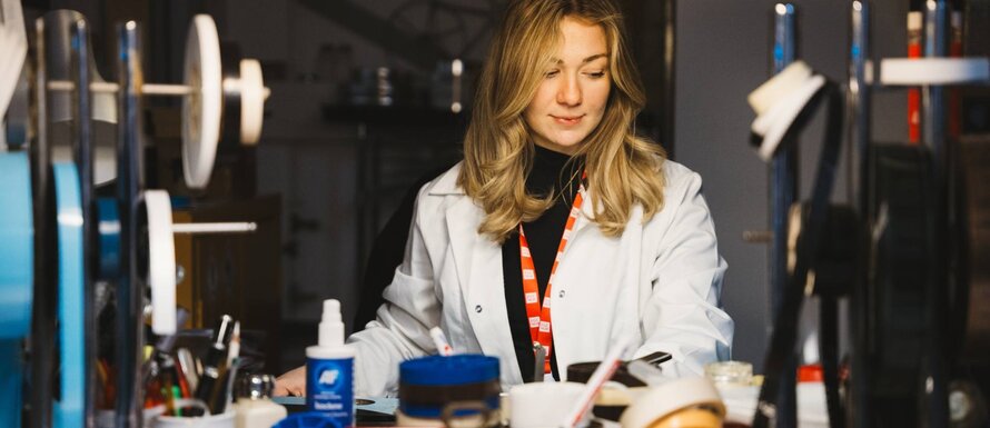 Angharad sits at a winding bench inspecting 16mm film. She's wearing a white lab coat.