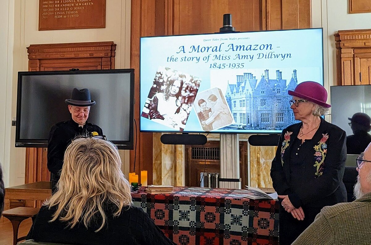 Two people giving a presentation to an audience during the 'Queer Tales from Wales' event in the Council Chamber at the National Library of Wales