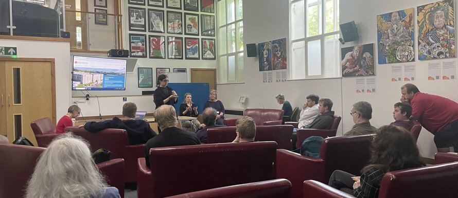 A wide view from the back of a room of people sitting in comfortable red sofas, looking toward the front, where a person is standing and presenting. There are two BSL interpreters facing the audience too. The walls are adorned with various posters and works of art.