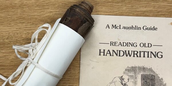 Image of a scroll wrapped in paper and tied with string, next to the cover of a volume bearing the title 'Reading Old Handwriting'