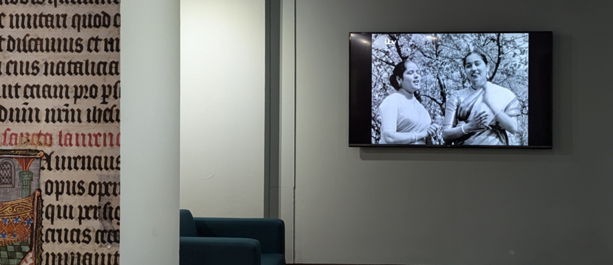 A photo of the large, wall-mounted screen, part of a comfortable chair just visible to the left of it. On the screen is a black and white image of two women in saris who are mid-song. An ITV logo is in the upper left corner of the screen. To the left of the image is part of a pillar covered in gothic script.