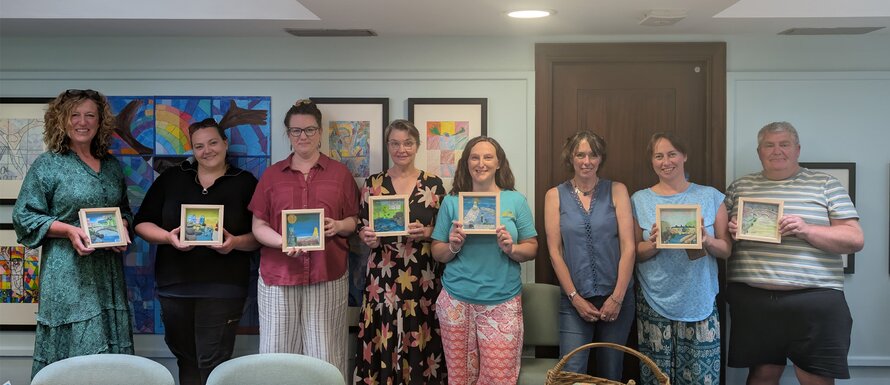 Eight participants stand indoors, each proudly displaying a framed painting they created during an art workshop. The room is bright and welcoming, with colorful artwork lining the walls and a table in the foreground scattered with paints, scissors, and other creative supplies—capturing the collaborative and expressive spirit of the event.