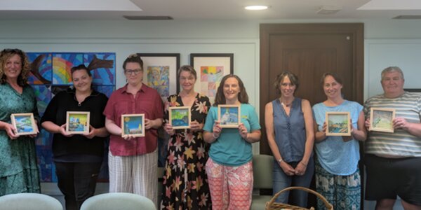 Eight participants stand indoors, each proudly displaying a framed painting they created during an art workshop. The room is bright and welcoming, with colorful artwork lining the walls and a table in the foreground scattered with paints, scissors, and other creative supplies—capturing the collaborative and expressive spirit of the event.