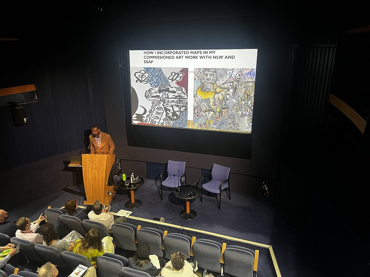 Mfikela Jean Samuel at a lectern in front of a screen showing details of two artworks