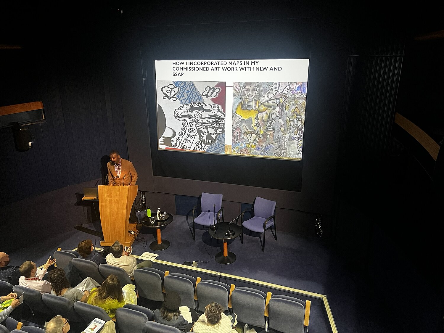 Mfikela Jean Samuel at a lectern in front of a screen showing details of two artworks