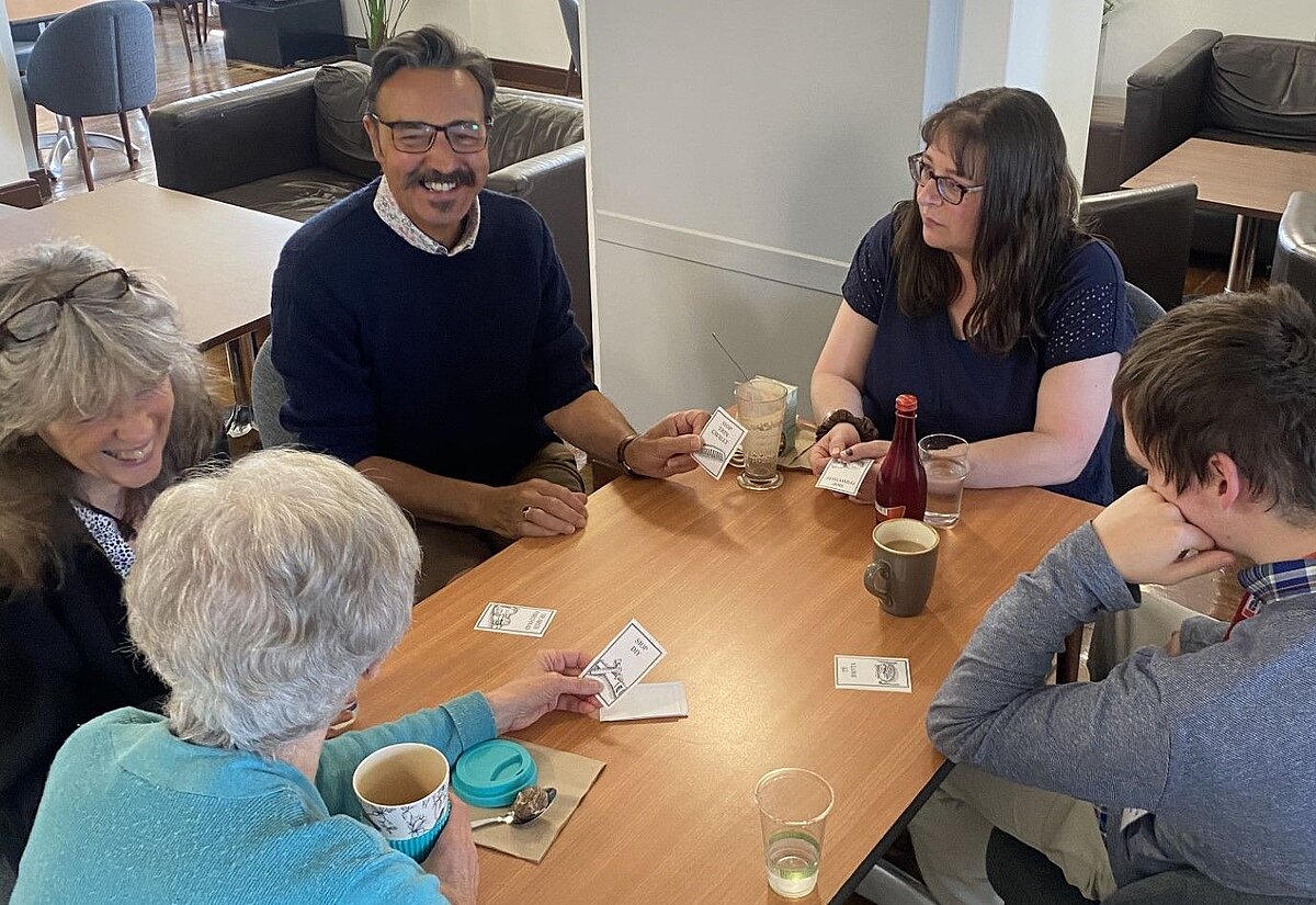 Five people sitting at a table in the Pen Dinas Cafe at the Library. They have hot drinks and cakes on the table