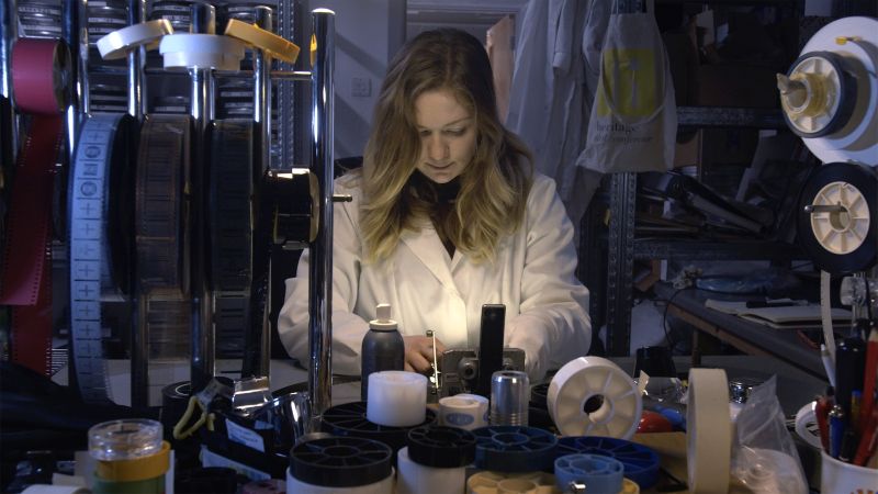 A wide-angle view of a woman wearing a white 'lab'-style coat sitting at a workbench inspecting film. She is surrounded by many reels of film and other equipment.