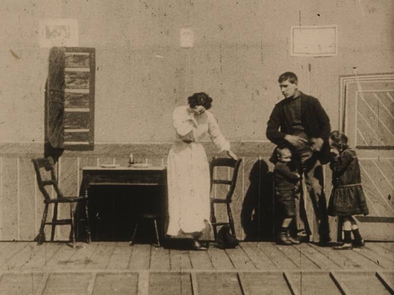 A screenshot from Haggar's The Sheep Stealer, showing a theatrical style dining room set. A woman stands near a table, her head in her hands, while a man looks on, two children holding on to his middle.